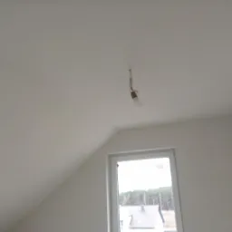 Gładka, pomalowana na biało skośna ściana poddasza z oknem, widoczna żarówka na kablu zwisająca z sufitu.