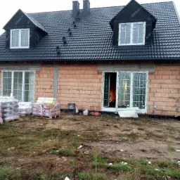 Dom w trakcie budowy z czarną dachówką, oknami PCV i stosem materiałów budowlanych na paletach, widoczne pustaki ceramiczne i folia dachowa Topdach