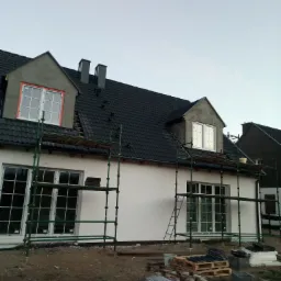 Nowy dom jednorodzinny w trakcie wykańczania elewacji, widoczne rusztowania, okna z pomarańczową folią zabezpieczającą, czarny dach z dachówki, przygotowane palety z materiałami budowlanymi.