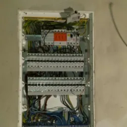 Rozdzielnica elektryczna z widocznymi bezpiecznikami, przewodami i elementami zabezpieczającymi, zamontowana w ścianie w stanie surowym.