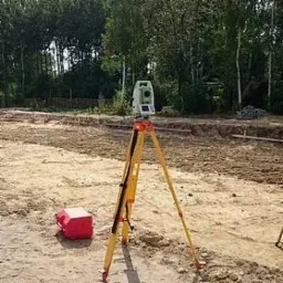 Żółty teodolit ustawiony na trójnogu na placu budowy z wykopem w tle, obok czerwona skrzynka narzędziowa.