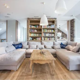 Jasny salon z dużym narożnikiem, drewnianym stolikiem kawowym i regałem z książkami, oświetlony wiszącymi lampami.