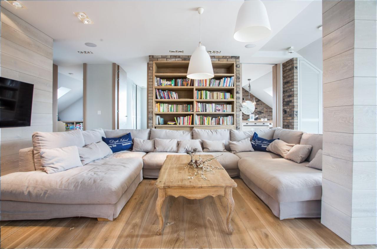 Jasny salon z dużym narożnikiem, drewnianym stolikiem kawowym i regałem z książkami, oświetlony wiszącymi lampami.