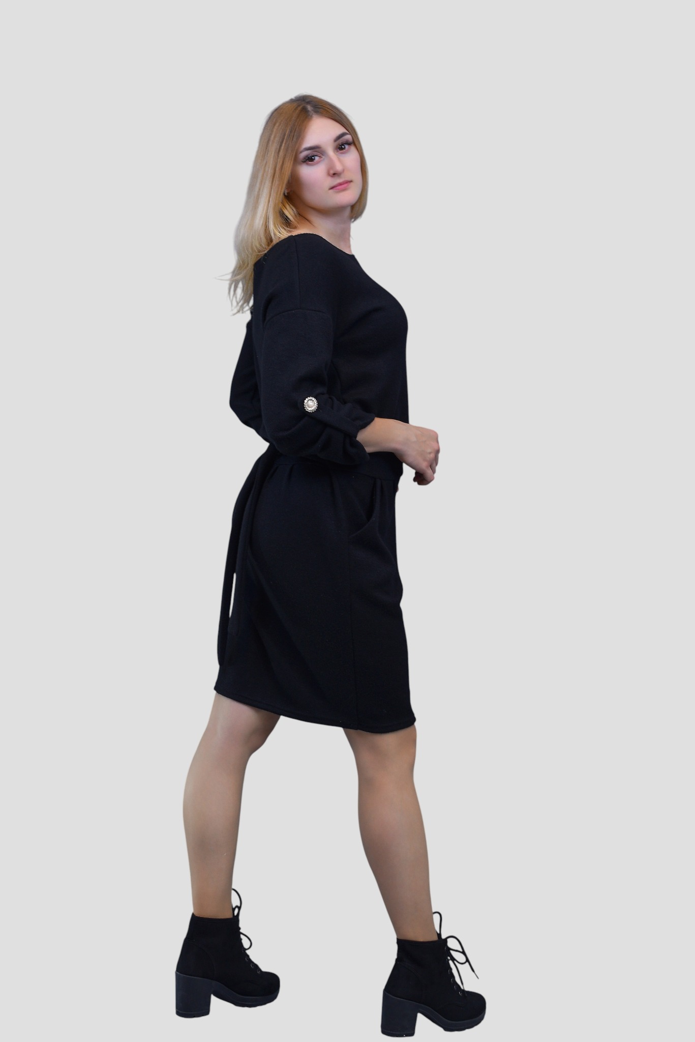 Kobieta prezentuje czarną sukienkę z wiązaniem w pasie i ozdobnymi guzikami na rękawach, pozując w czarnych botkach na obcasie na neutralnym tle.