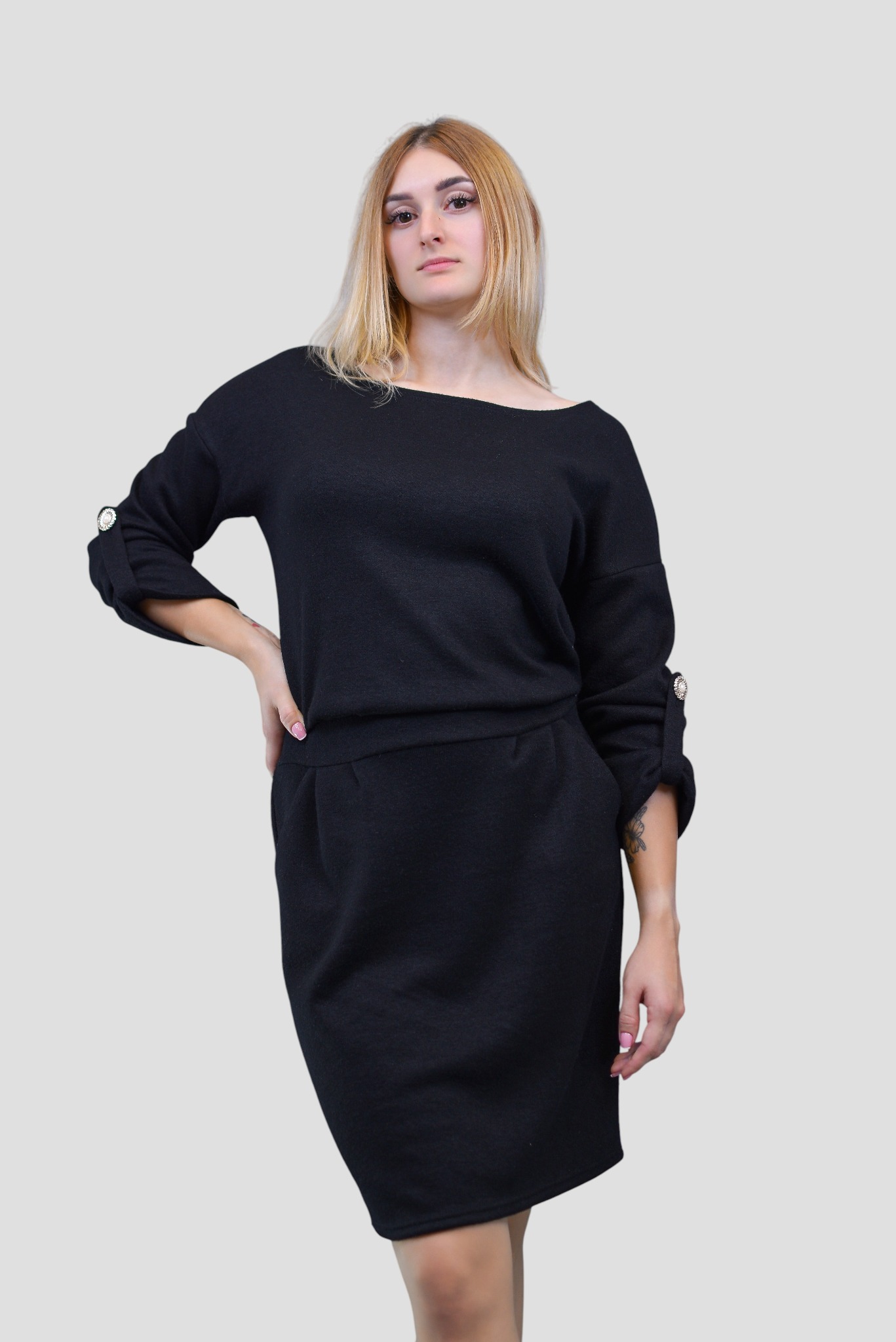 Kobieta w czarnej sukience z rękawami 3/4, zdobionymi ozdobnymi guzikami, pozuje na jasnym tle. Sukienka o prostym kroju, długości do kolan.