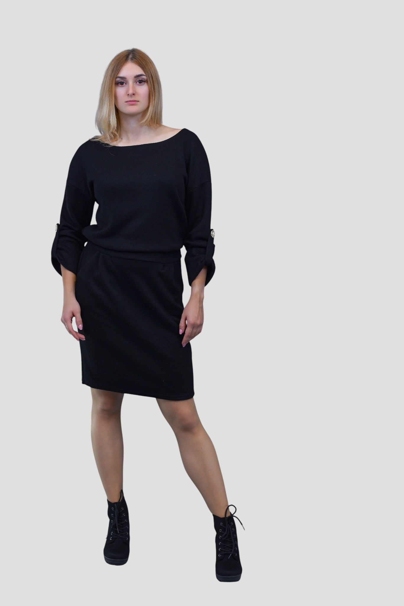 Kobieta prezentuje czarną, dzianinową sukienkę mini z długim rękawem i mankietami zapinanymi na guziki, do której dobrała czarne sznurowane botki na platformie.