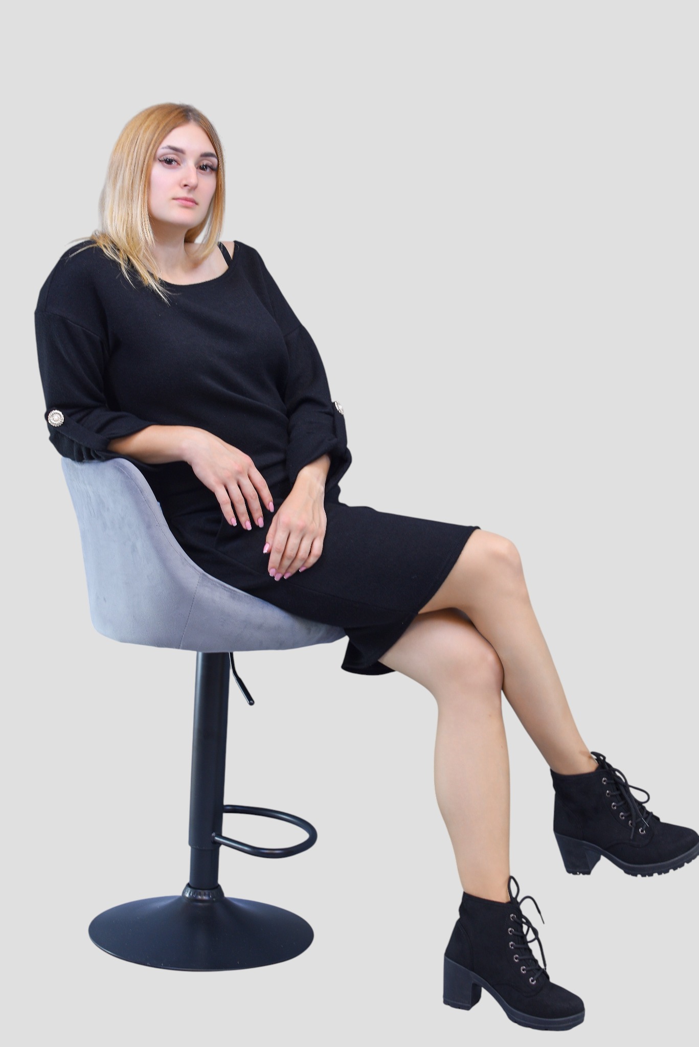 Kobieta w czarnej sukience siedzi na szarym krześle barowym, prezentując strój i buty. Neutralne tło, oświetlenie studyjne, widoczne detale sukienki i obuwia.