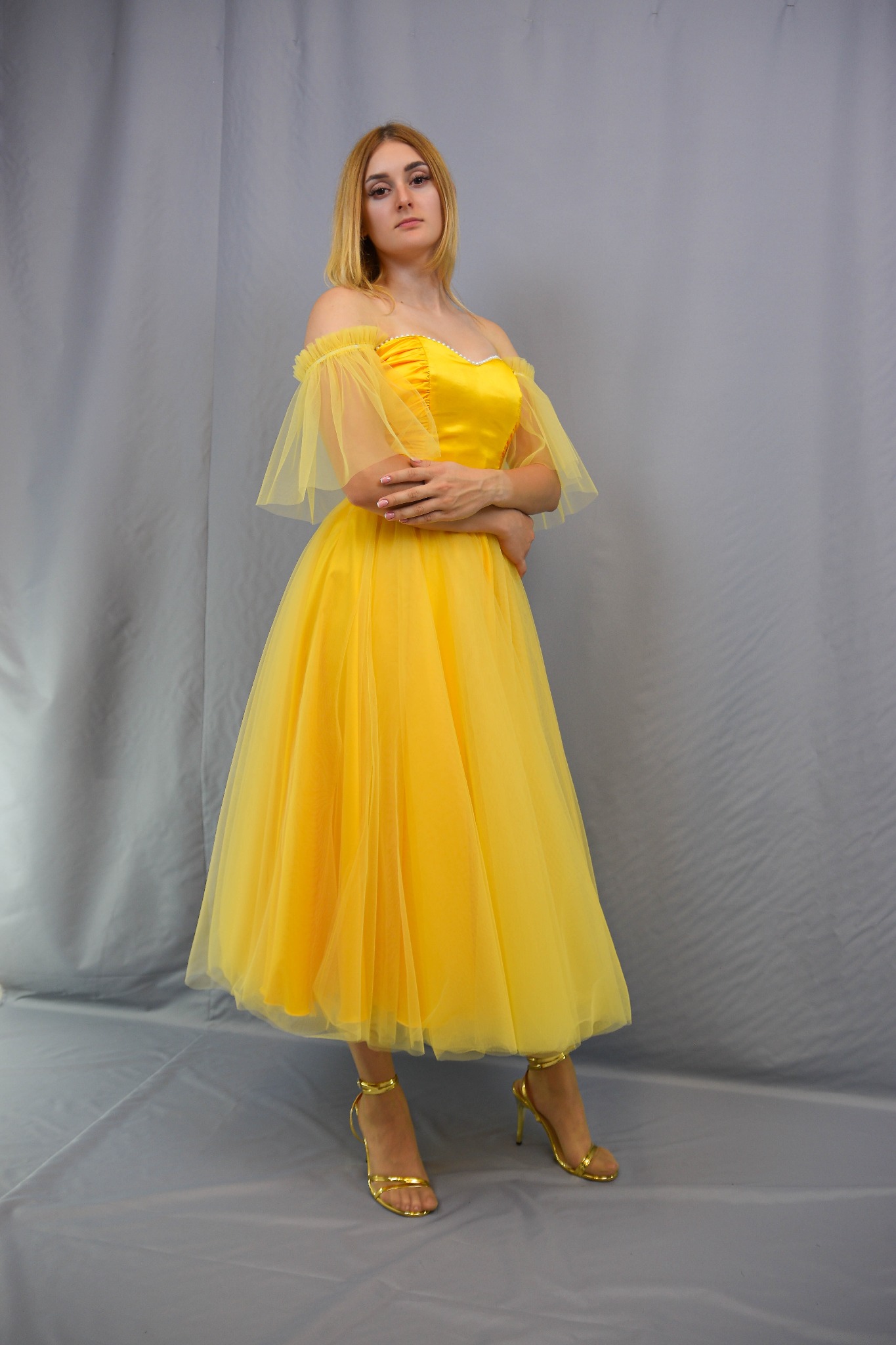 Kobieta w żółtej balowej sukni z tiulu i złotych sandałach na obcasie, prezentuje kreację od krawcowej z Gdańska na tle szarej tkaniny.