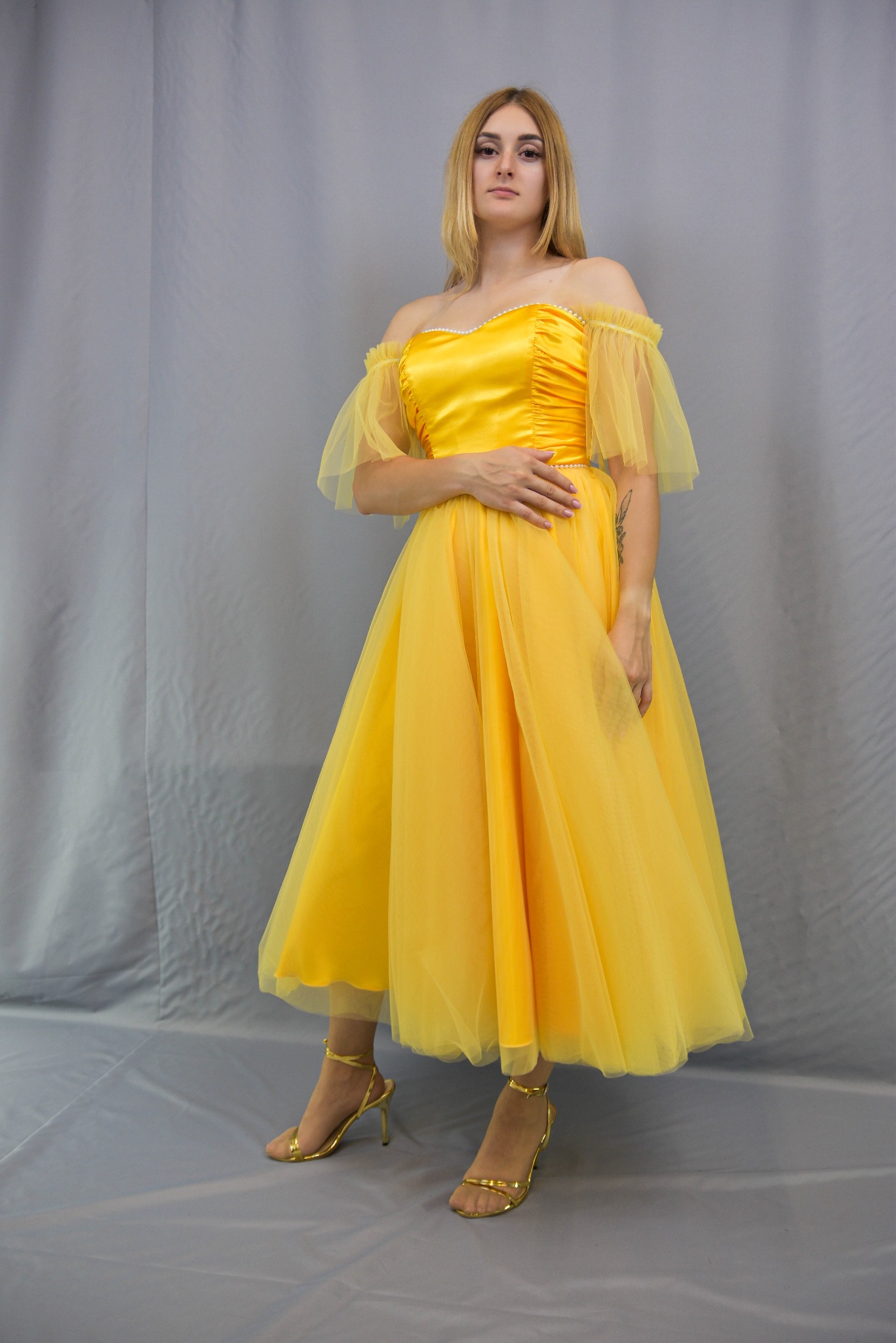 Kobieta w żółtej, tiulowej sukience z odsłoniętymi ramionami i złotych sandałach na obcasie, pozuje na szarym tle. Sukienka ma drapowany gorset i bufiaste rękawki.
