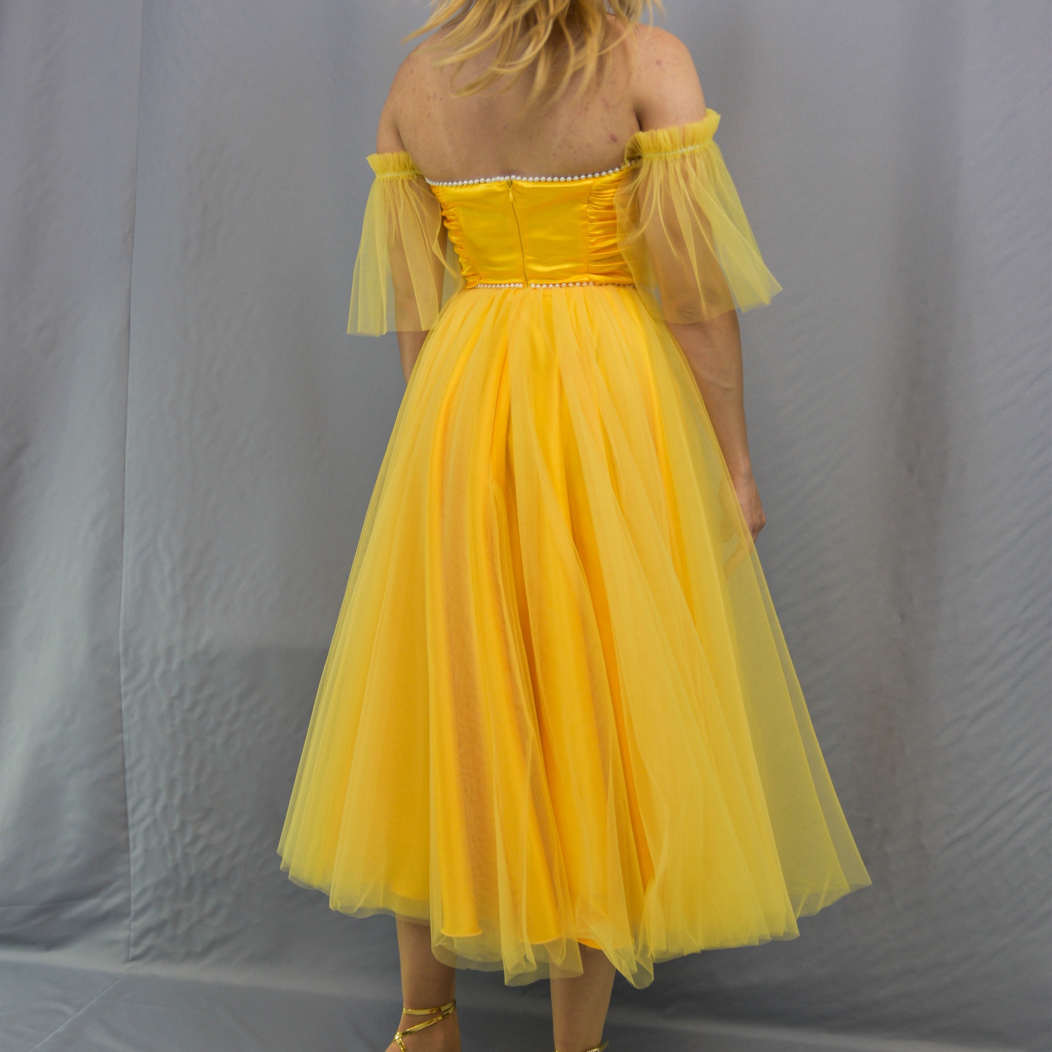 Żółta, tiulowa sukienka z odkrytymi ramionami, ozdobiona perełkami, widziana od tyłu na tle szarego materiału. Widoczne złote sandałki.