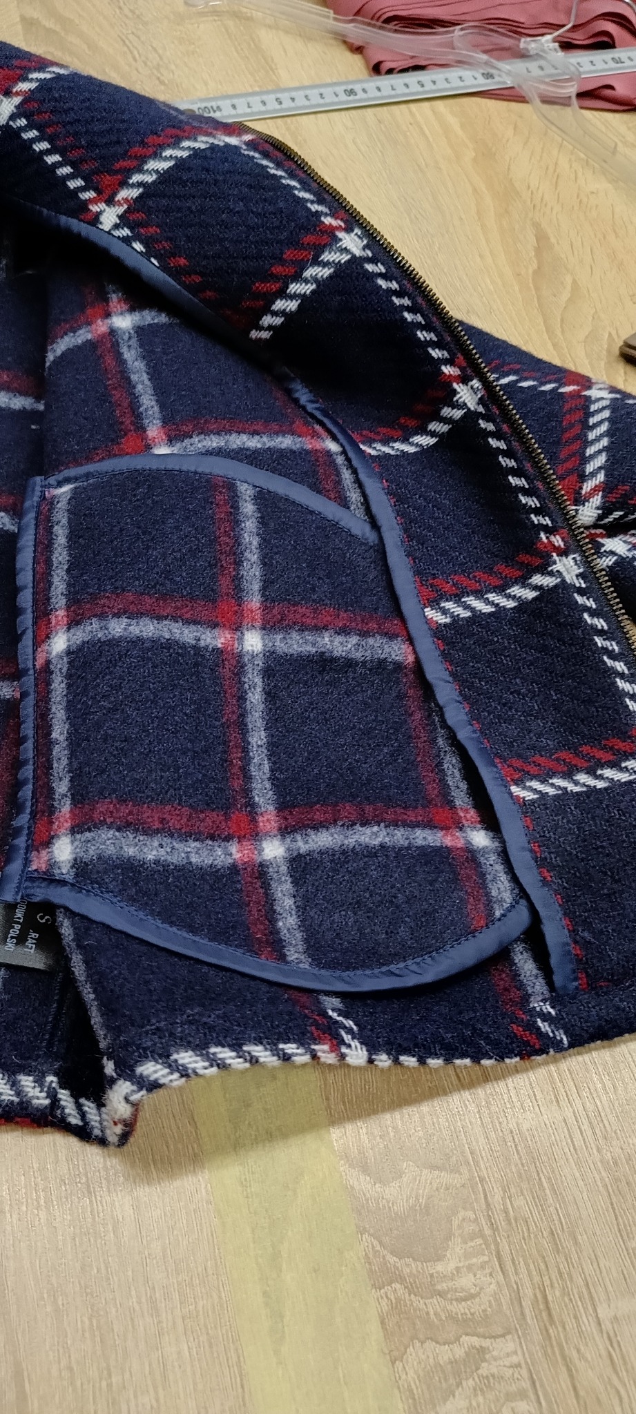 Fragment uszytej, wełnianej kurtki w kratę, z kieszenią i zamkiem błyskawicznym, leżącej na drewnianym stole. Widoczny rozmiar S na metce.
