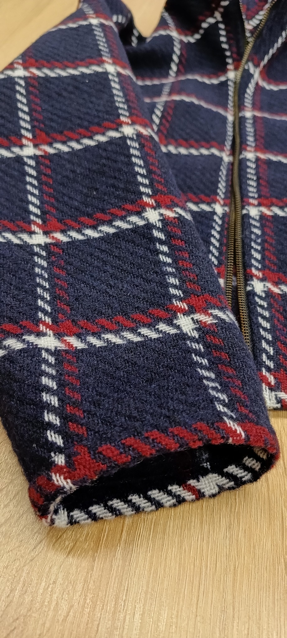 Fragment granatowej kurtki w czerwoną i białą kratę z metalowym zamkiem błyskawicznym. Widoczny mankiet z wykończeniem w biało-czarne paski.