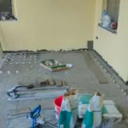 Balkon w trakcie układania płytek podłogowych, widoczne krzyżyki dystansowe, paczki z klejem, przecinarka do płytek oraz fragmenty płytek w jasnym kolorze. Ściany pomalowane na jasnoż&oacute;łty kolor.