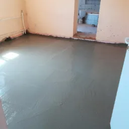 Świeżo wylana, mokra wylewka samopoziomująca w pomieszczeniu z widocznymi rurami grzewczymi przy ścianie i otwartym przejściem do wykończonej łazienki z białymi płytkami.