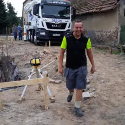 Uśmiechnięty pracownik w roboczych butach idzie po placu budowy fundamentów, w tle betoniarka firmy Walbet i poziomica laserowa.