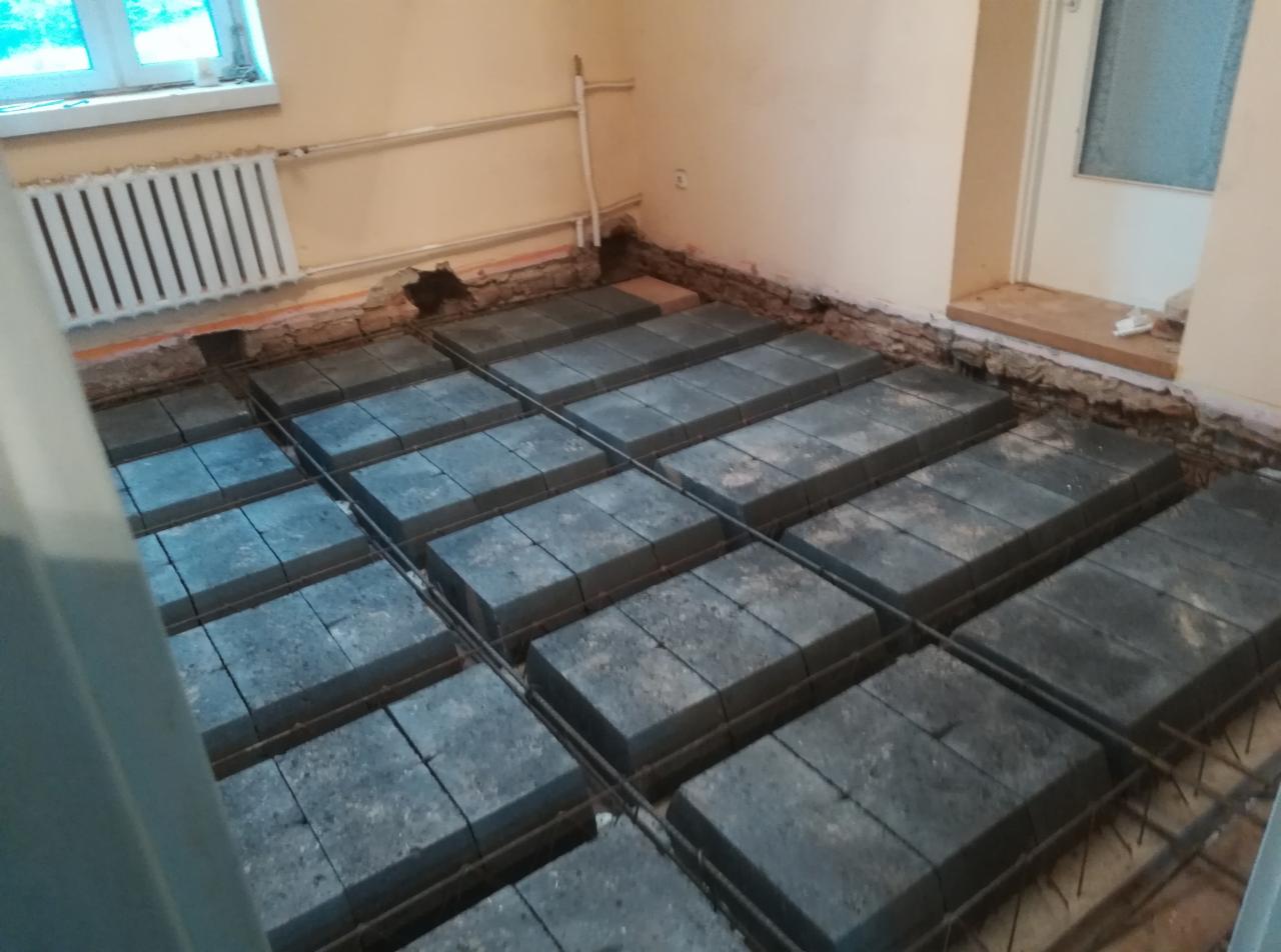 Przygotowanie podłoża pod wylewkę: ułożone bloczki betonowe na siatce zbrojeniowej w pomieszczeniu z widocznym grzejnikiem i fragmentem ściany z ubytkami tynku.