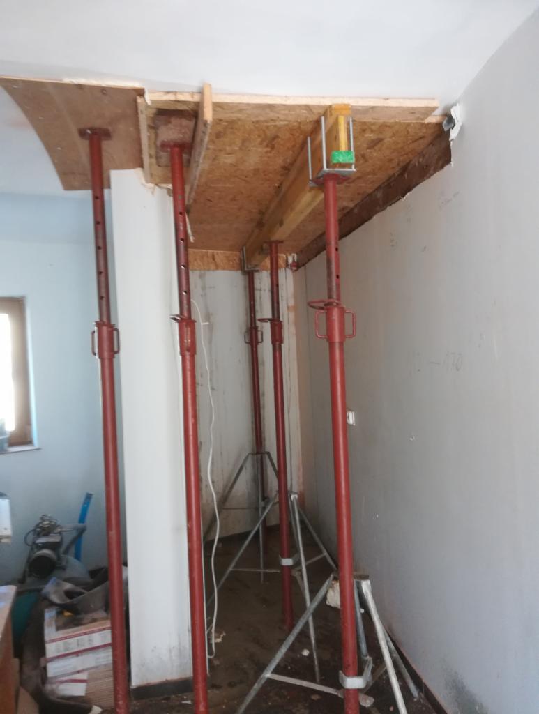 Wnętrze w trakcie remontu z widocznymi czerwonymi stemplami podtrzymującymi drewnianą konstrukcję sufitu, częściowo odsłonięta instalacja elektryczna, ściany z widocznymi śladami po pracach...