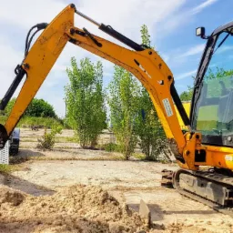 Żółto-czarna mini koparka gąsienicowa na placu budowy, z łyżką zanurzoną w piasku, częściowo przykrytą czerwoną tkaniną, w tle drzewa i przyczepa.