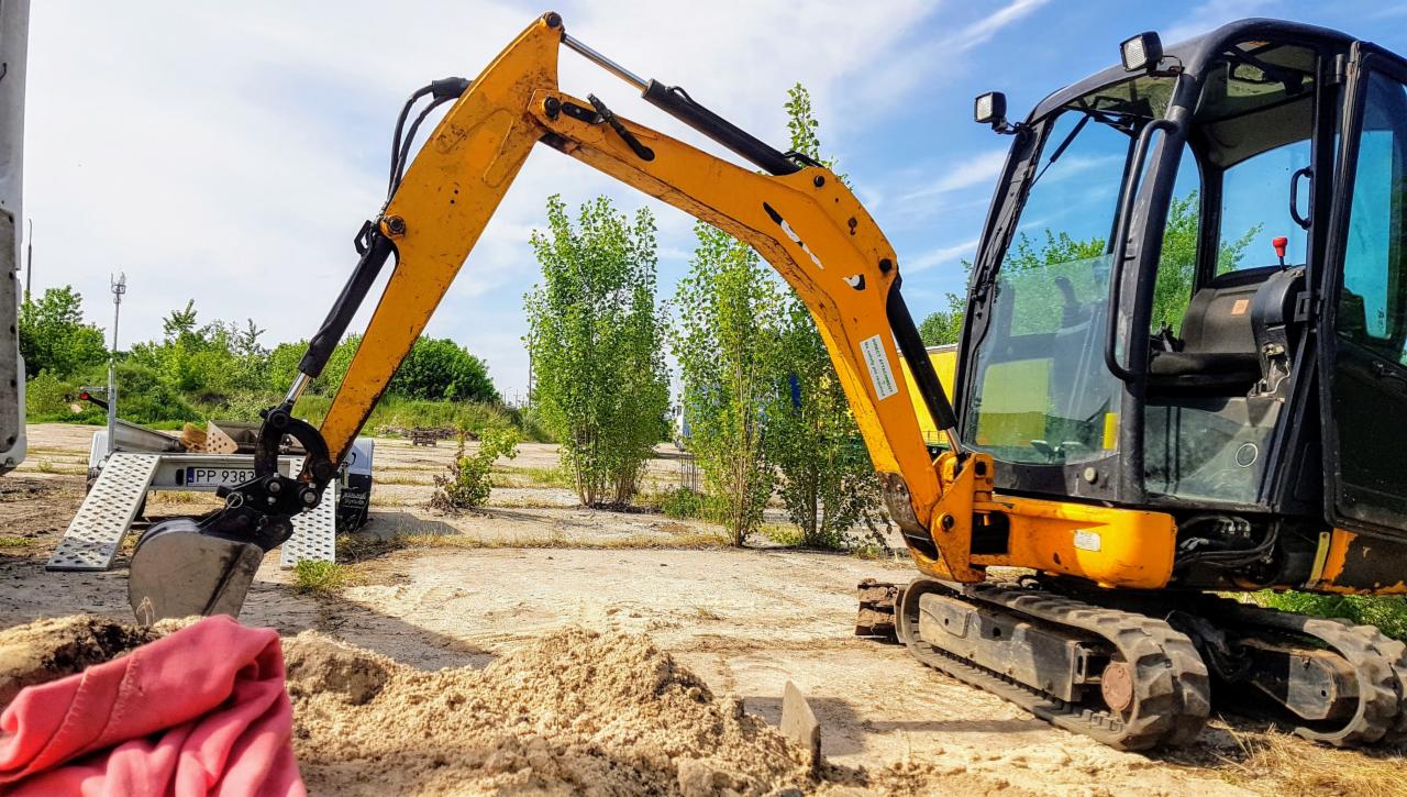 Żółto-czarna mini koparka gąsienicowa na placu budowy, z łyżką zanurzoną w piasku, częściowo przykrytą czerwoną tkaniną, w tle drzewa i przyczepa.