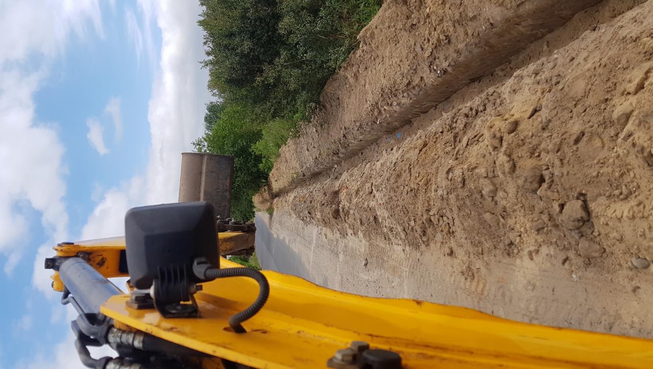 Żółta koparka wykopuje rów w ziemi, widok z perspektywy operatora, w tle drzewa i błękitne niebo z chmurami.