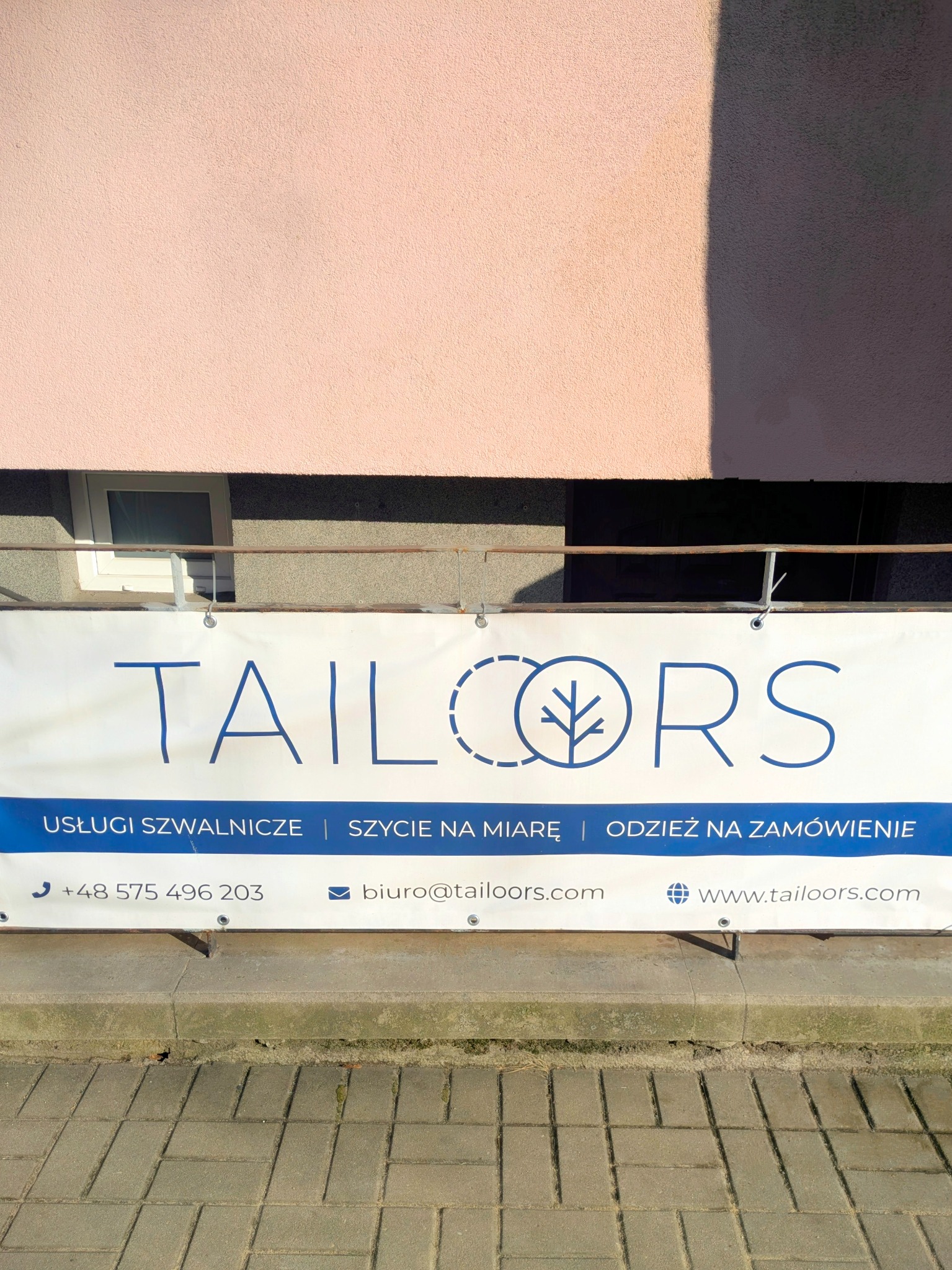 Baner reklamowy firmy Tailors oferującej usługi szwalnicze i szycie na miarę, zawieszony na tle różowej ściany budynku, z widocznym adresem e-mail i stroną internetową.