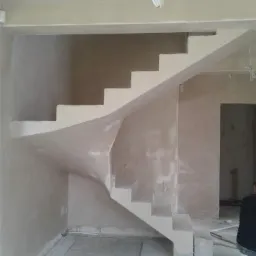 Wnętrze w trakcie prac wykończeniowych: spiralne schody z surowego betonu pokryte świeżo nałożonym tynkiem, widoczne ślady po pracach na ścianach i podłodze, zwisające przewody elektryczne.