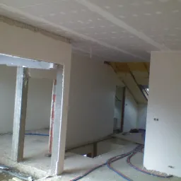 Wnętrze w trakcie remontu: sufit wykończony płytami gipsowo-kartonowymi, widoczne profile i łączenia, ściany częściowo przygotowane, instalacje elektryczne na podłodze, otwór w stropie.