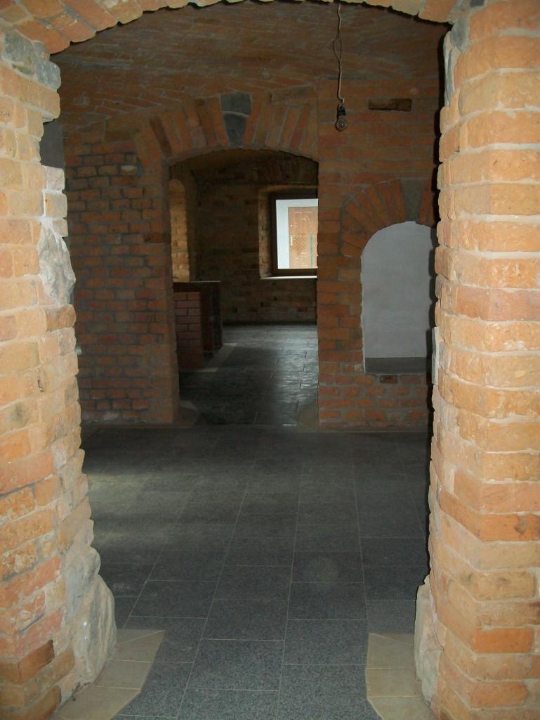 Wnętrze piwnicy z ceglanymi łukami i szarą, kwadratową podłogą z płytek, widok przez ceglane przejście z surowymi ścianami.