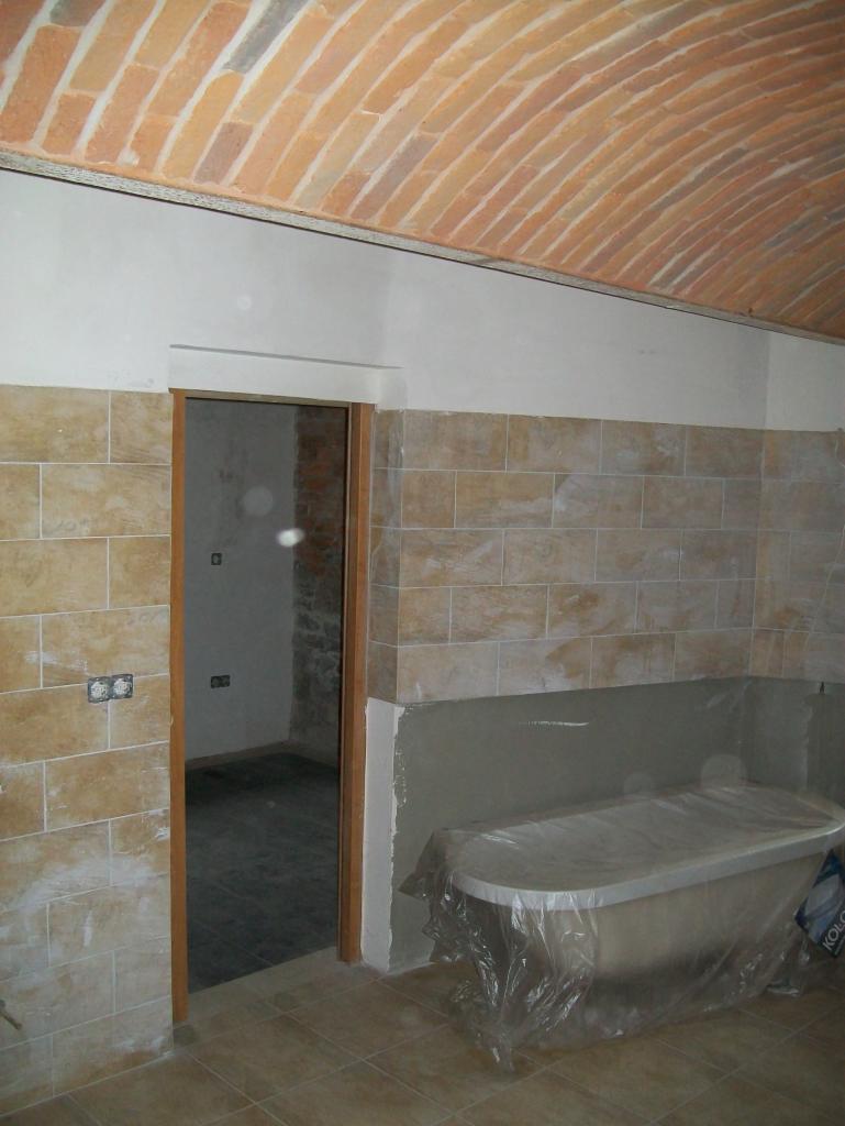 Łazienka w trakcie remontu: beżowe płytki na ścianach, wanna zabezpieczona folią, ceglany łuk na suficie, widoczne puszki elektryczne bez osprzętu.