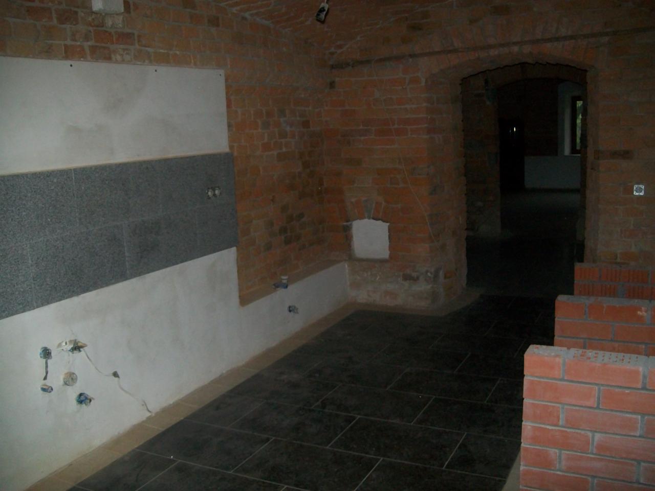 Wnętrze w trakcie remontu: fragment ściany z położoną częścią szarej glazury, surowa cegła, ciemna podłoga z płytek, widoczne instalacje elektryczne i hydrauliczne.