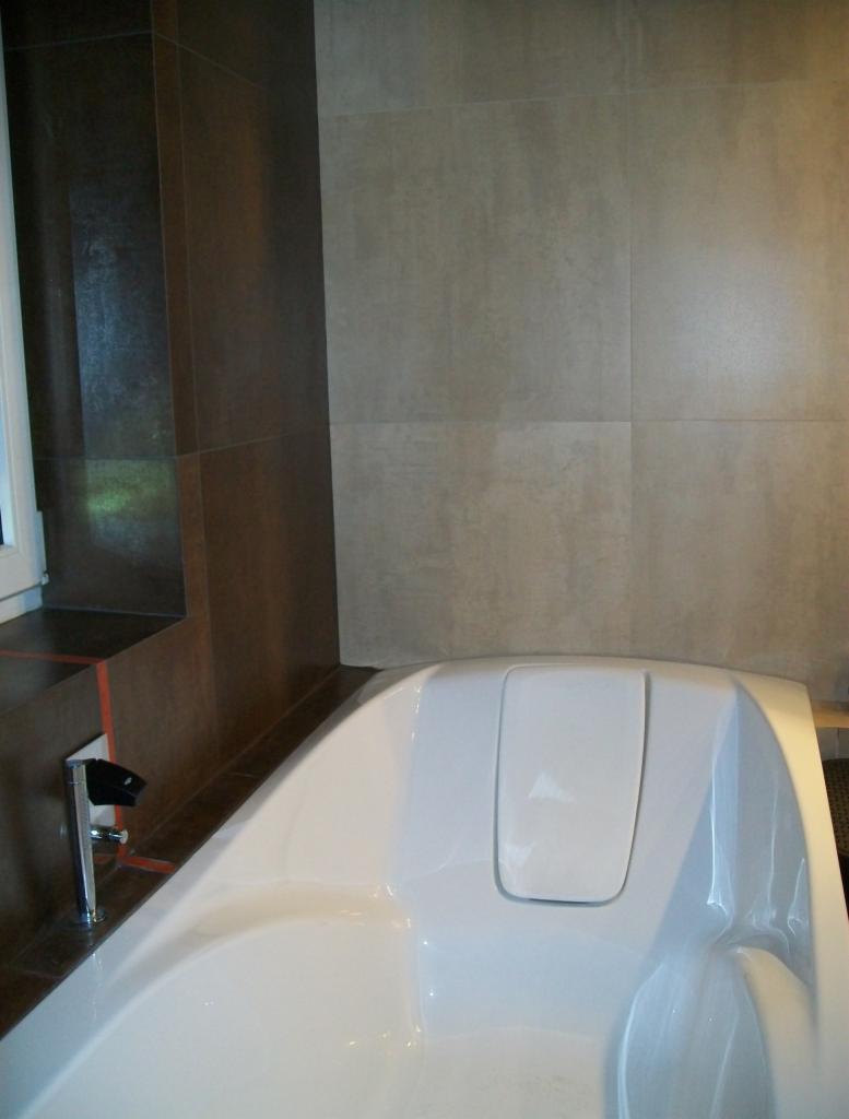 Nowoczesna, biała wanna z chromowaną baterią, przylegająca do ściany wyłożonej beżowymi, kwadratowymi płytkami ceramicznymi o matowym wykończeniu i ciemnobrązowej ściany przy oknie.