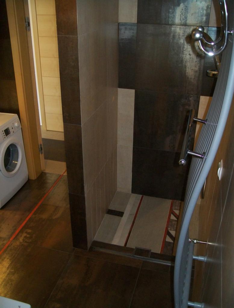 Nowoczesna kabina prysznicowa z ciemnymi, metalizowanymi płytkami w odcieniach brązu i szarości, z elementami dekoracyjnymi w podłodze, widoczny grzejnik drabinkowy i pralka w tle.