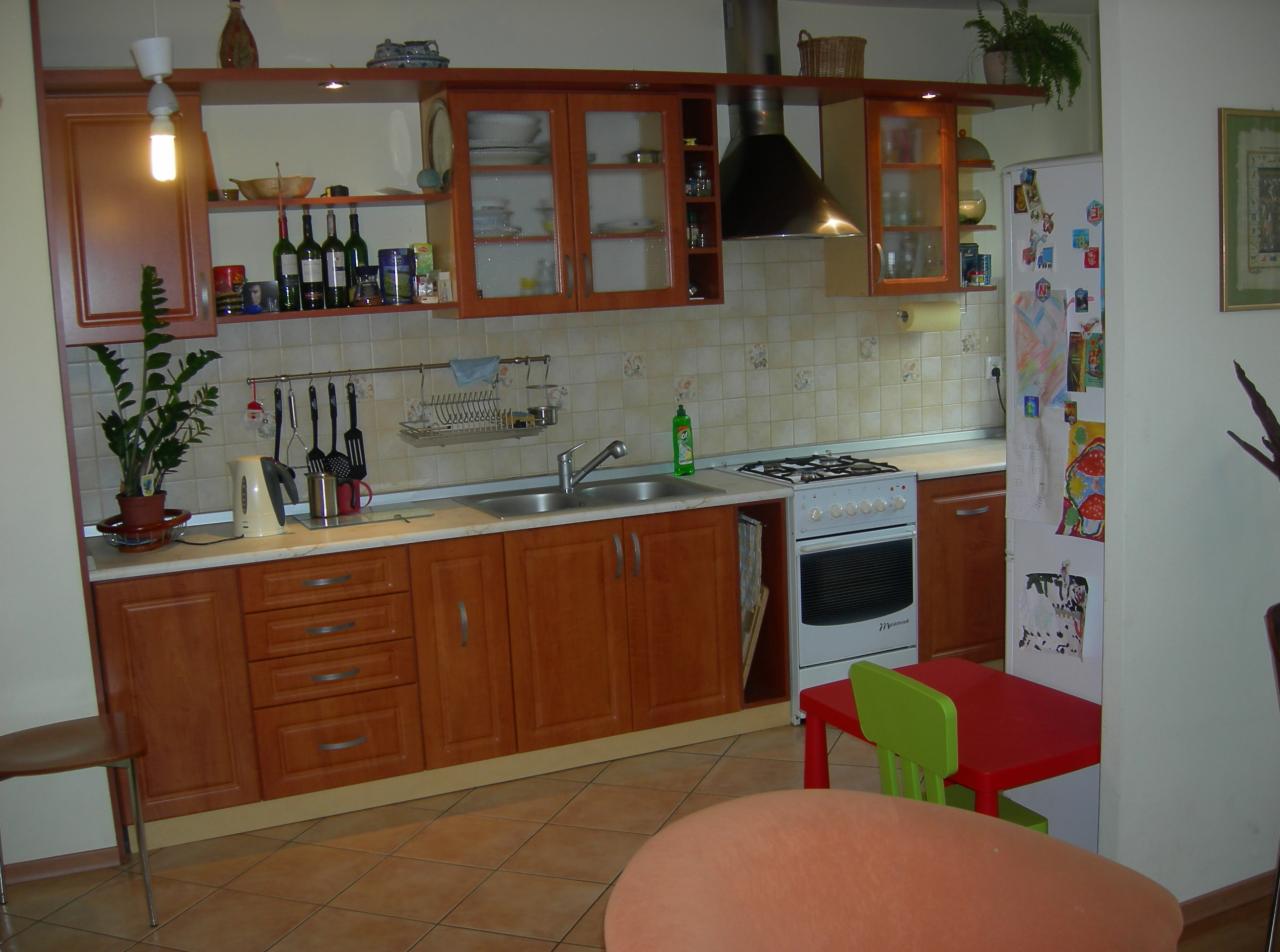 Kuchnia z drewnianymi szafkami, białą kuchenką gazową i płytkami ściennymi w beżowym odcieniu. Na blacie stoją akcesoria kuchenne, a na półce nad zlewem znajdują się butelki. Przy ścianie stoi...