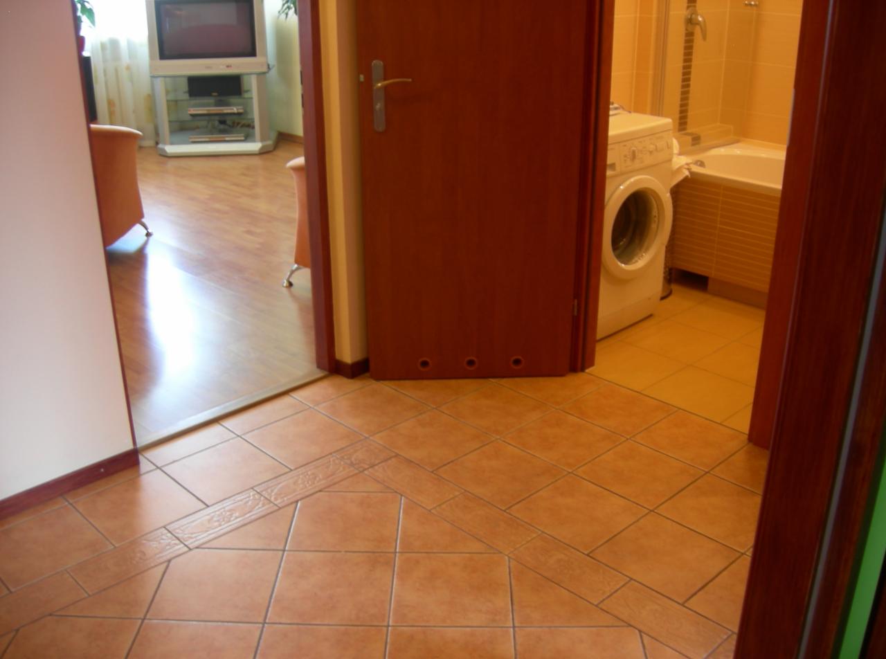 Beżowe płytki podłogowe ułożone we wzór z dekoracyjnym pasem, widok na łazienkę z pralką i wanną oraz fragment salonu z parkietem, przez otwarte drzwi.