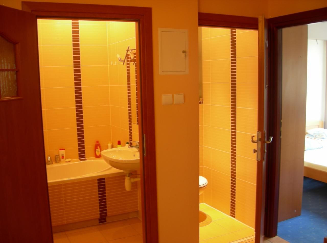 Widok przez otwarte drzwi na łazienkę z wanną obudowaną płytkami, umywalką i toaletą, wykończoną w odcieniach pomarańczy i brązu, z widocznym fragmentem sypialni w tle.