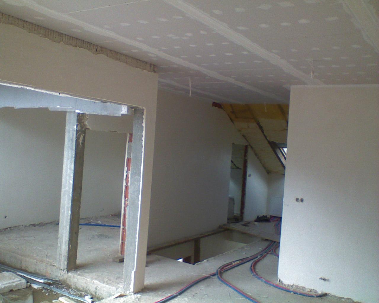 Wnętrze w trakcie remontu: sufit wykończony płytami gipsowo-kartonowymi, widoczne profile i łączenia, ściany częściowo przygotowane, instalacje elektryczne na podłodze, otwór w stropie.