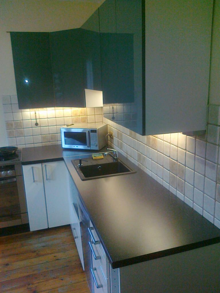 Nowoczesna kuchnia z ciemnym blatem, kwadratowymi płytkami na ścianie i drewnianą podłogą; widoczne oświetlenie podszafkowe.