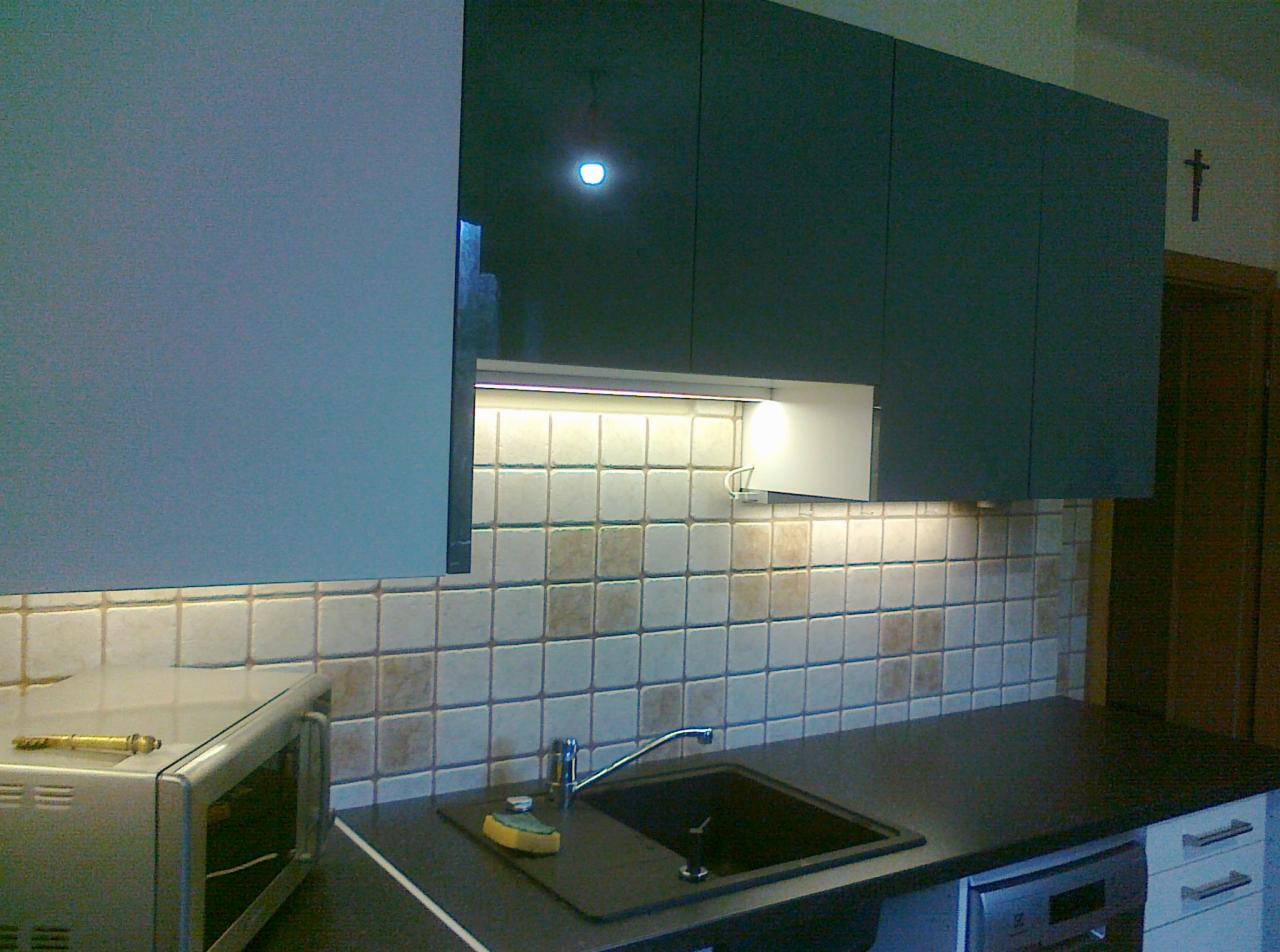 Fragment kuchni z ciemnymi szafkami wiszącymi, podświetlanymi od dołu, oraz jasnymi, kwadratowymi płytkami na ścianie nad blatem, gdzie widoczny jest zlew i bateria.