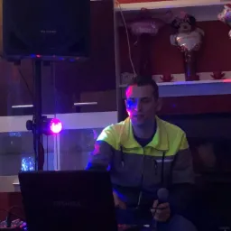 Mężczyzna w kurtce z mikrofonem i laptopem marki Toshiba, obok głośnik, oświetlony fioletowym światłem w pomieszczeniu z czerwonymi ścianami i półką z dekoracjami.