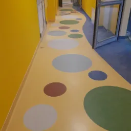 Korytarz w przedszkolu z żółtą wykładziną z kolorowymi, okrągłymi wzorami i żółtymi ścianami, widok z perspektywy.