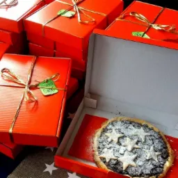 Stos czerwonych pudełek z ciastami, przewiązanych złotym sznurkiem i ozdobionych zielonymi etykietami, z otwartym pudełkiem prezentującym ciasto ze śliwkami i gwiazdkami z cukru pudru.