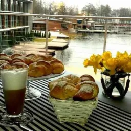 Śniadanie na tarasie z widokiem na jezioro: rogaliki posypane cukrem pudrem na paterze i w koszyku, kawa latte w szklance, żółte narcyzy w wazonie, czarno-biały bieżnik.