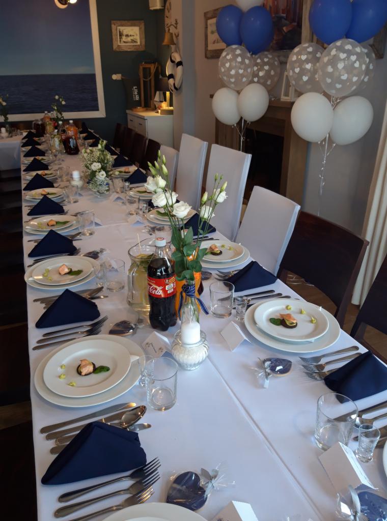 Długi stół nakryty białym obrusem, z dekoracjami kwiatowymi w wazonach, granatowymi serwetkami, talerzami z przystawkami, napojami i balonami w tle.
