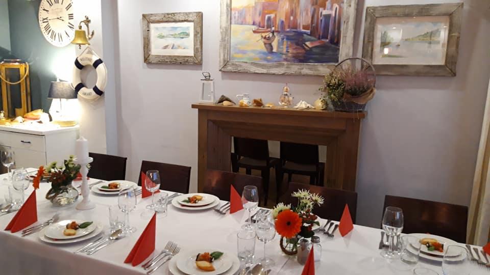 Uroczysty stół nakryty białym obrusem, z czerwonymi serwetkami, kwiatami i zastawą, w tle kominek i dekoracje marynistyczne, sugerujące aranżację tematyczną.