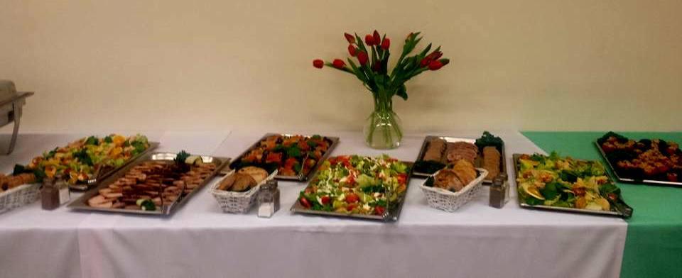 Stół cateringowy z różnorodnymi sałatkami, wędlinami i pieczywem, udekorowany wazonem z czerwonymi tulipanami, ustawiony na białym obrusie z fragmentem zielonego obrusu z prawej strony.
