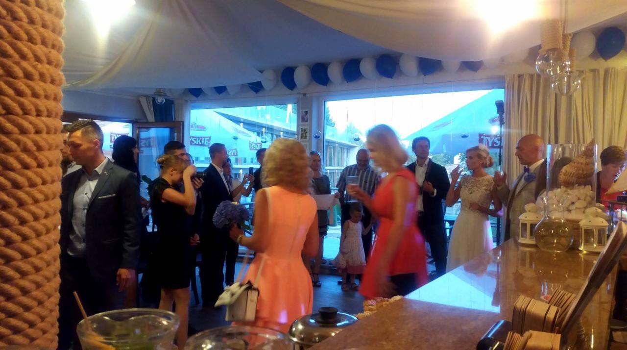 Uroczystość w lokalu: goście w strojach wieczorowych, panna młoda w sukni ślubnej, dekoracje balonowe i szklane naczynia na barze.