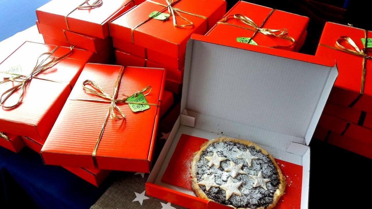 Stos czerwonych pudełek z ciastami, przewiązanych złotym sznurkiem i ozdobionych zielonymi etykietami, z otwartym pudełkiem prezentującym ciasto ze śliwkami i gwiazdkami z cukru pudru.