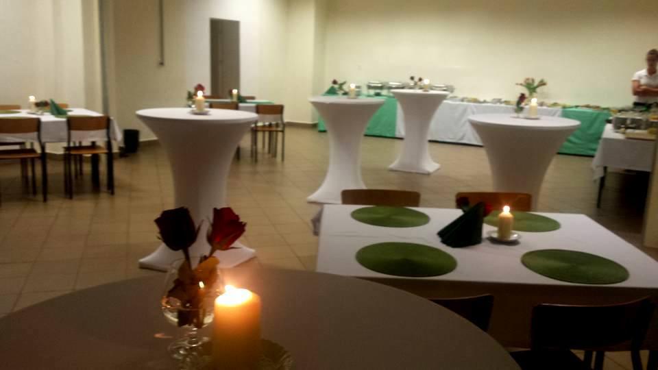 Sala bankietowa z nakrytymi stołami, białe obrusy, dekoracje kwiatowe w kieliszkach i świece na stołach, bufet w tle, przygotowana na przyjęcie.