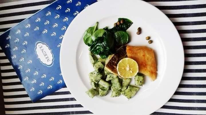 Elegancko podane danie rybne na białym talerzu: smażony filet, zielone gnocchi, świeży szpinak, kapary i plasterek cytryny. W tle menu w marynarskim stylu na czarno-białym, pasiastym obrusie.