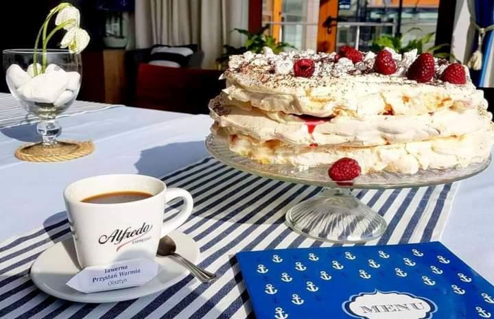 Trzypiętrowy tort bezowy z malinami na szklanej paterze, filiżanka kawy Alfredo Espresso, menu w niebieskiej okładce z motywem kotwic na stole w biało-niebieskie pasy.